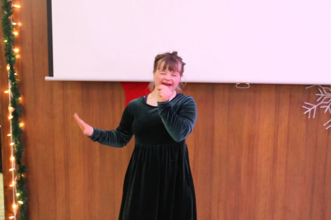 A young girl in a black dress laughing and covering her mouth, standing in front of a wooden wall with holiday decorations.