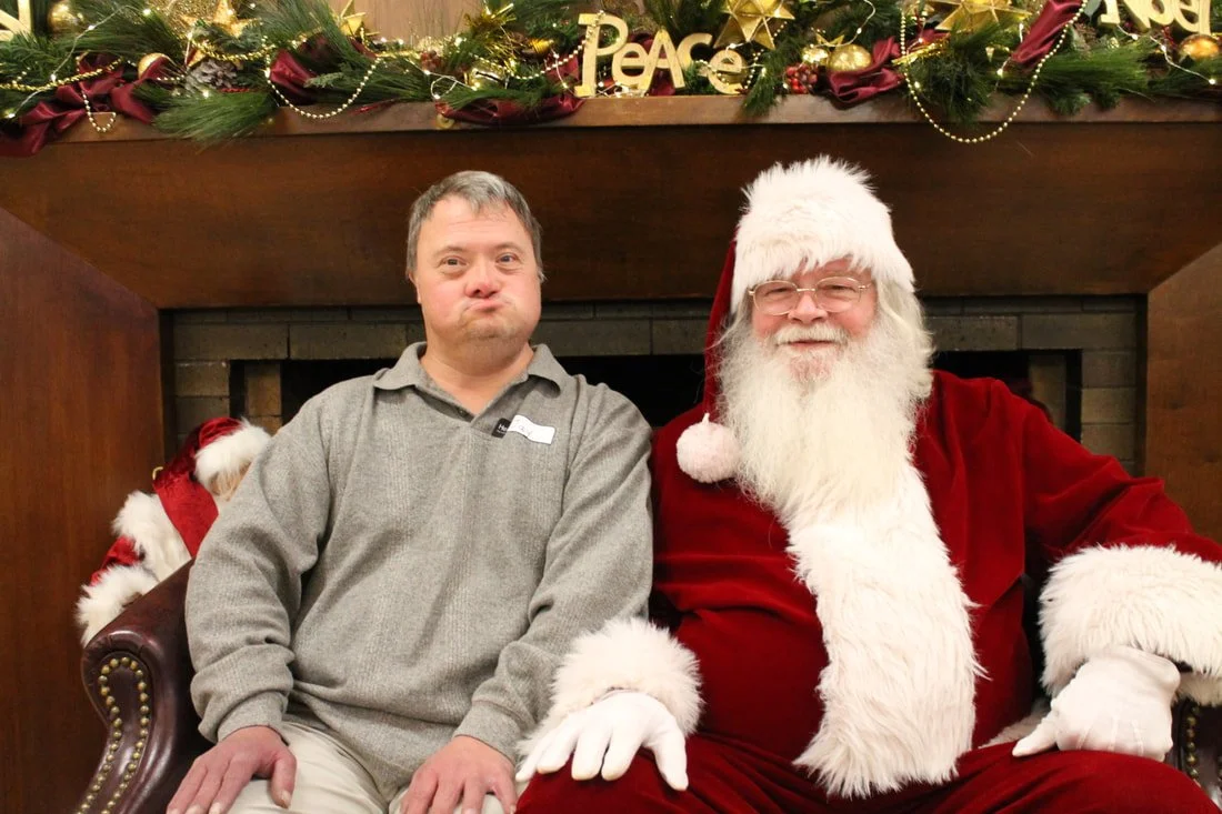 A man sitting next to a person dressed as Santa Claus, both sitting on a chair in front of a decorated fireplace with holiday wreath and garland.