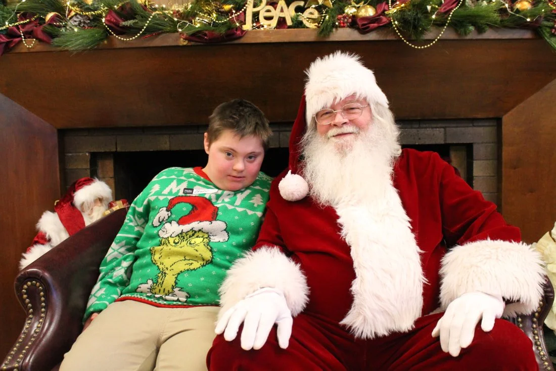 A boy sitting next to Santa Claus during a Christmas event, with holiday decorations including greenery, ornaments, and a sign that says 'Peace' in the background.
