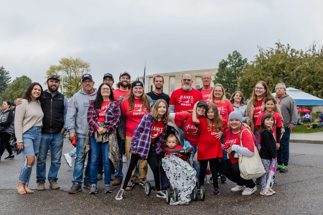 Group of adults and children gathered outdoors for a group photo, some wearing matching red shirts that say 'Alaina's Admirers'.