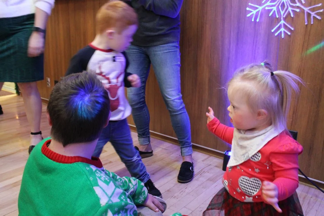 Young children and adults dancing and socializing at a holiday gathering with a blue neon snowflake on the wall.
