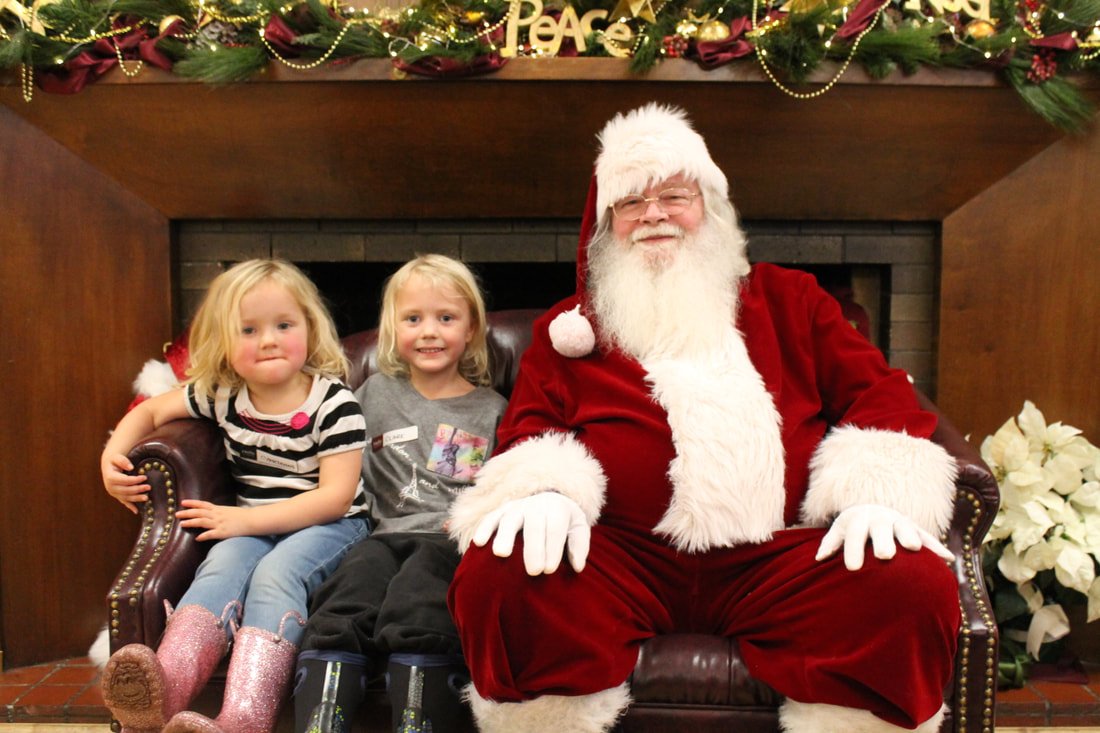 Two young girls sitting next to Santa Claus, who is dressed in a red suit with white trim, in a decorated fireplace setting with holiday ornaments and Christmas flowers.