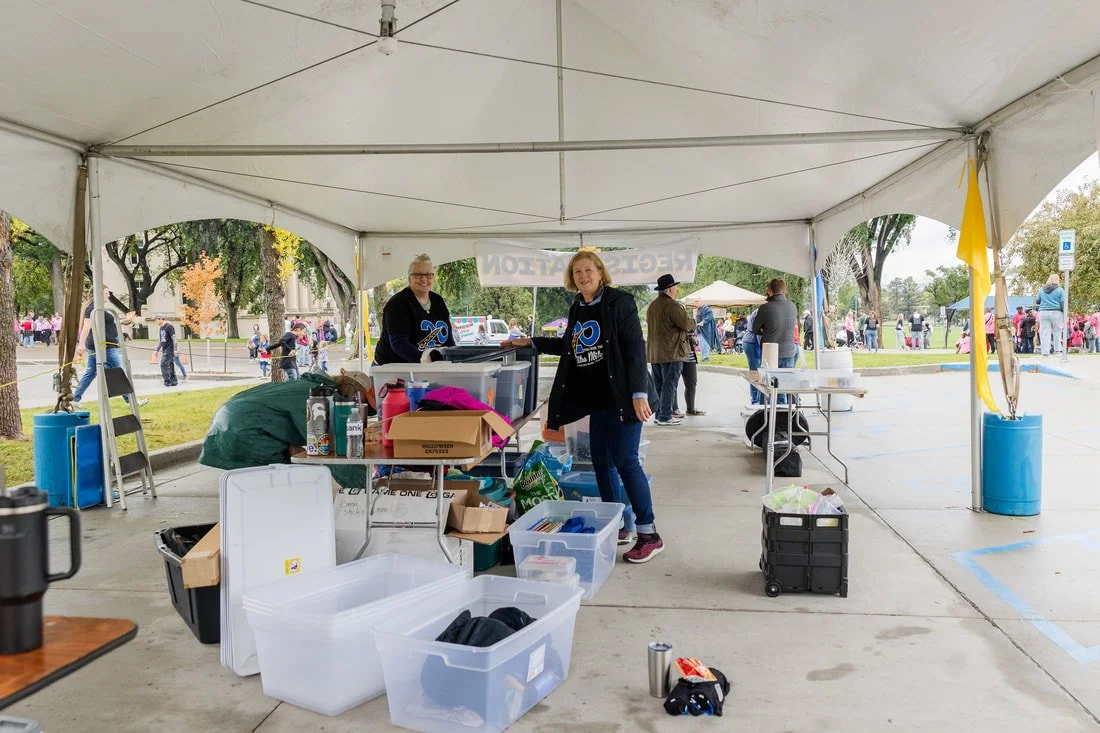 Two women standing under a white event tent at an outdoor park, with tables, storage bins, and supplies, surrounded by people and trees.
