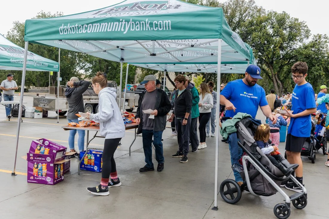 People gathered under a teal canopy tent with 'dakotacommunitybank.com' printed on it, participating in an outdoor event with food and activities.
