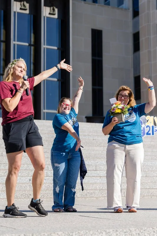 Three women standing on steps, one holding a microphone and a bouquet of flowers, celebrating outdoors near a large building.