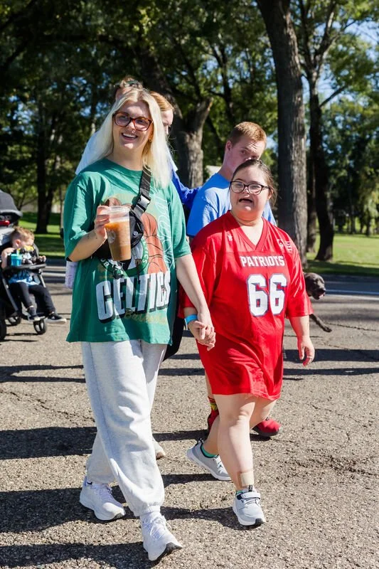 Group of smiling people walking outdoors in a park, wearing casual clothes, with trees and a street in the background.