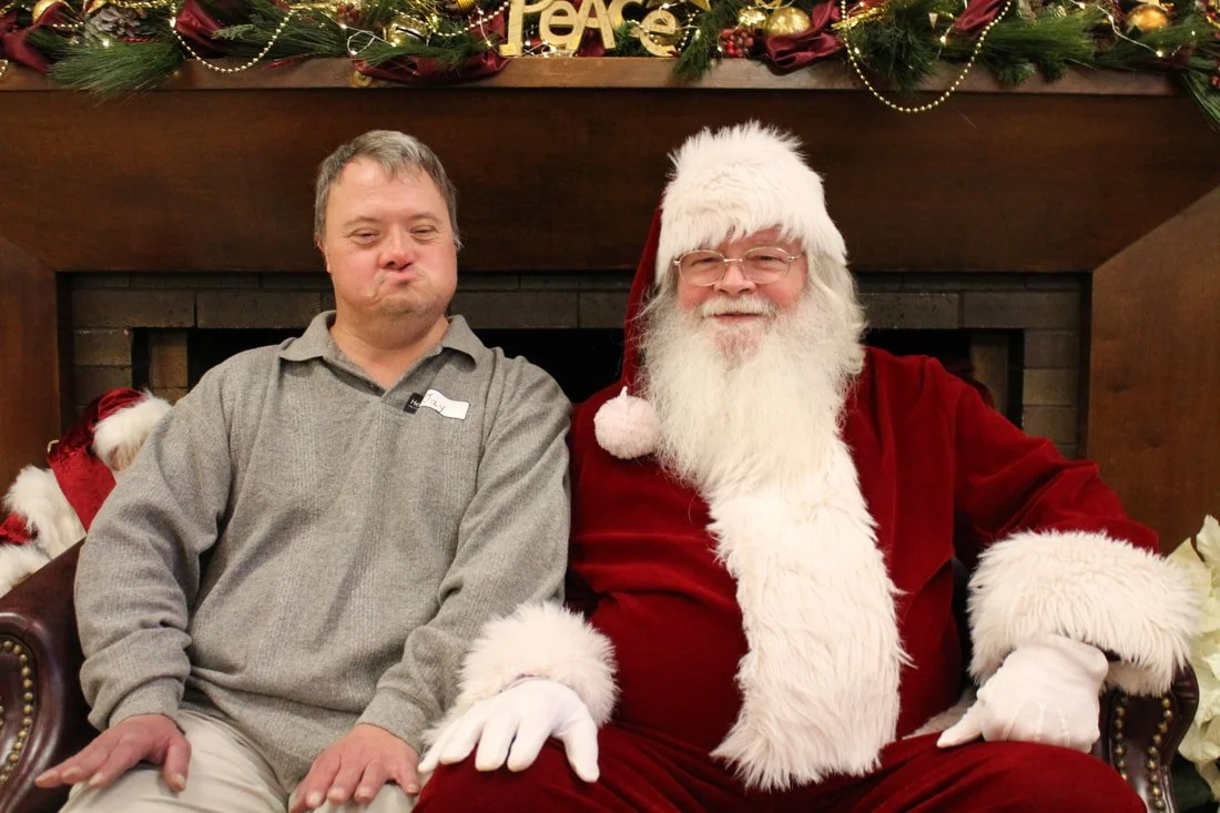 A man sitting next to a man dressed as Santa Claus, both smiling, in front of a Christmas fireplace with holiday decorations.
