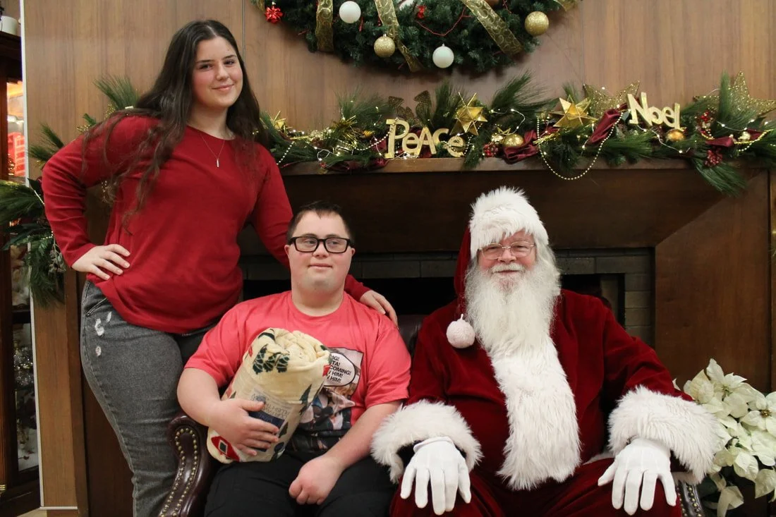 A woman and a young man sitting with Santa Claus in front of a Christmas decorated fireplace, with Christmas garlands, ornaments, and a wreath with a sign that says 'Peace' and 'Noel'.