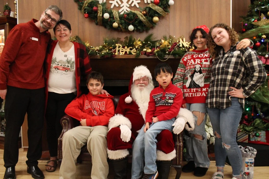 People grouped around Santa Claus in front of a Christmas tree and holiday decorations.