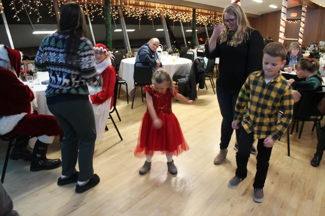 People dancing at a Christmas party, including children and adults, with festive holiday decorations and lights in the background.