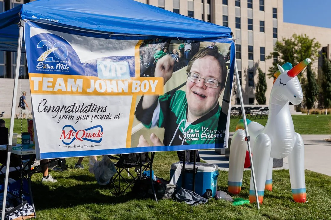 A canopy with a banner that reads 'Team John Boy' and 'Congratulations from your friends at McQuade Distributing Co., Inc.'. The banner features a large photo of a smiling young man with glasses and brown hair, wearing a green shirt, participating in