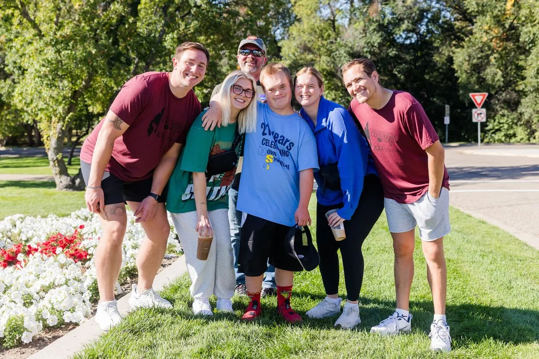 A group of six people smiling outdoors in a park, standing on grass near flowers and trees.