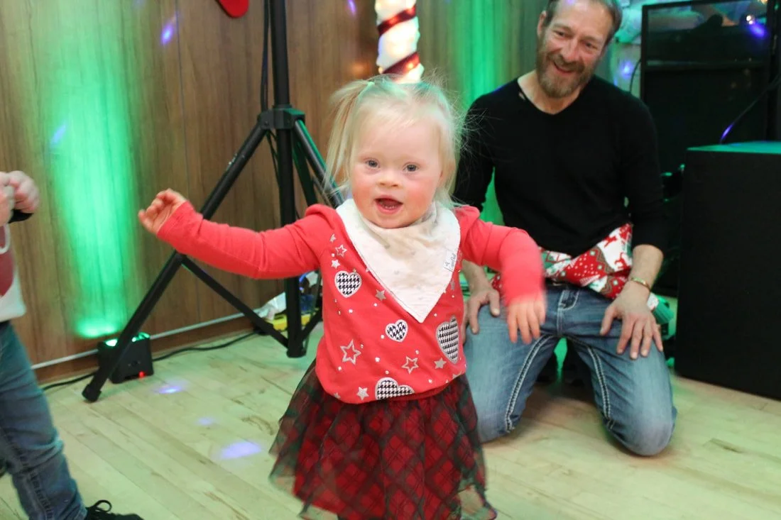 A young girl dancing at a holiday party, with a man sitting behind her on the floor, smiling. The girl is wearing a red holiday sweater with heart and star patterns, a tartan skirt, and a white bandana. The background features colorful lights and hol