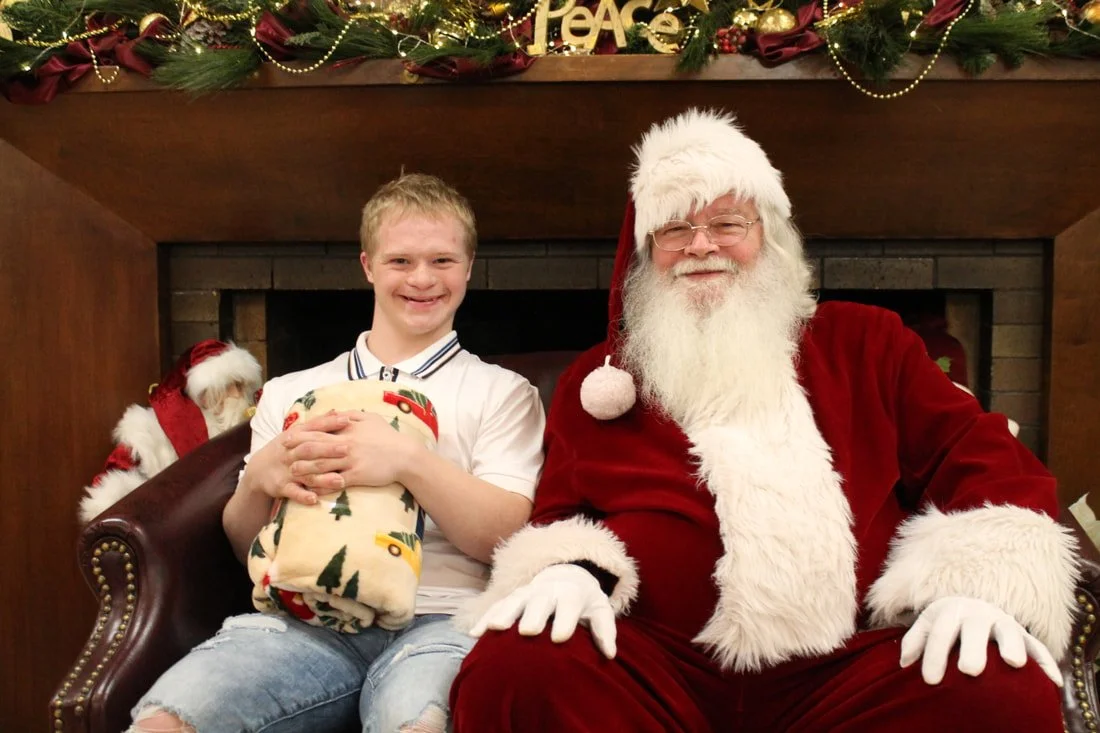 A young boy sitting on Santa Claus's lap, holding a wrapped Christmas gift, in front of a fireplace decorated with Christmas garland, ornaments, and a 