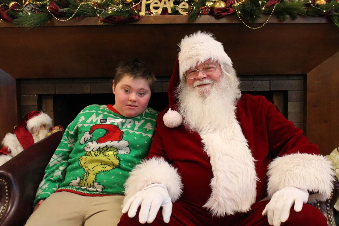 A young boy with a Christmas sweater sitting next to Santa Claus in front of a fireplace decorated for Christmas.