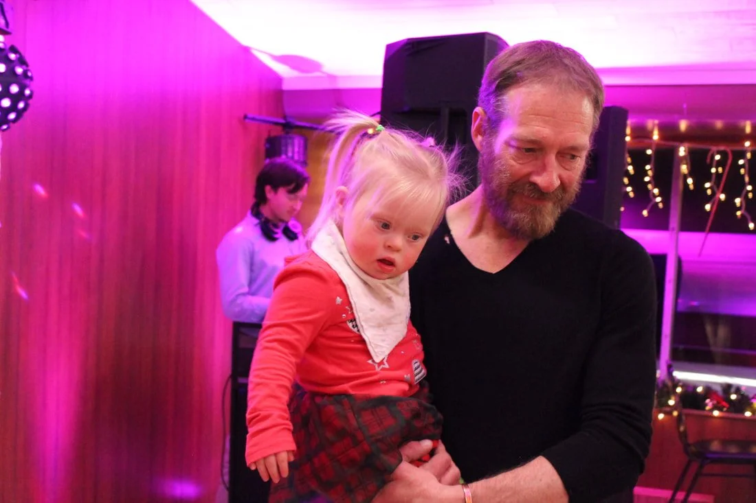 A man holding a young girl at a party or celebration with purple and pink lighting, decorations, and a person in the background with DJ equipment.
