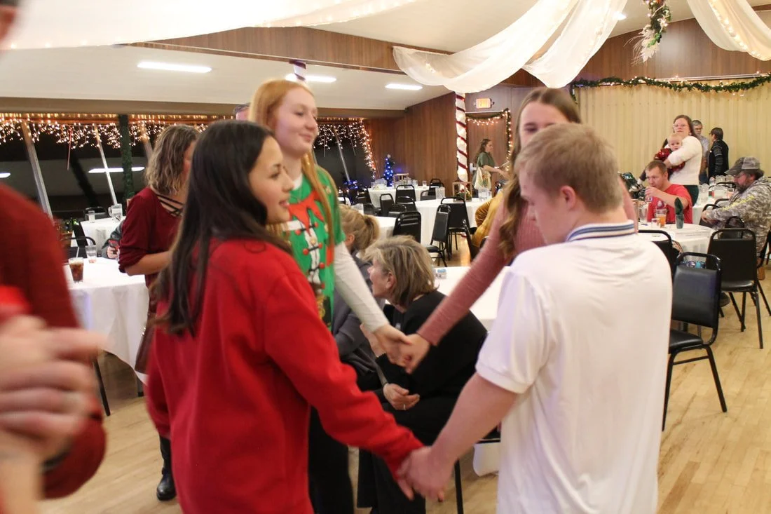 A group of children holding hands in the center of a decorated banquet hall with Christmas decorations, tables, and chairs.