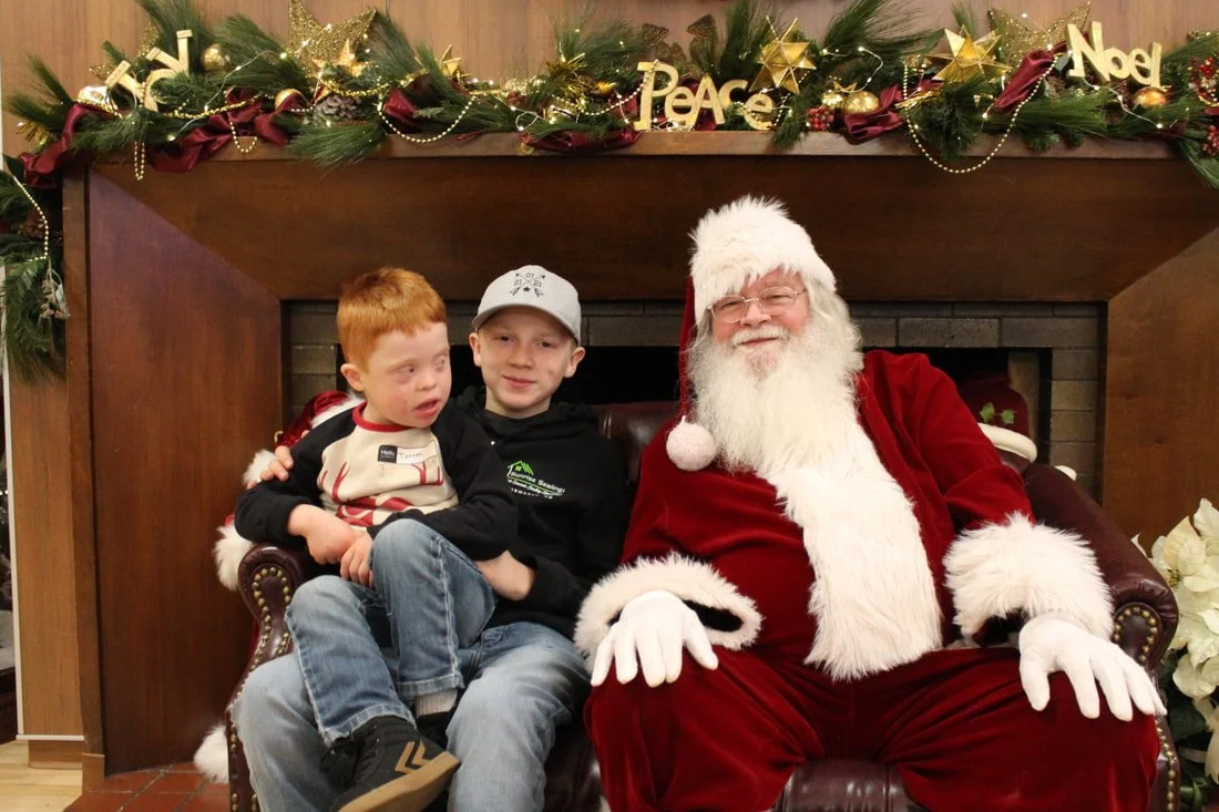 Two children sitting with Santa Claus in front of a decorated fireplace with Christmas garland, gold ornaments, and signs that say 'Peace,' 'Noel,' and 'Joy'.