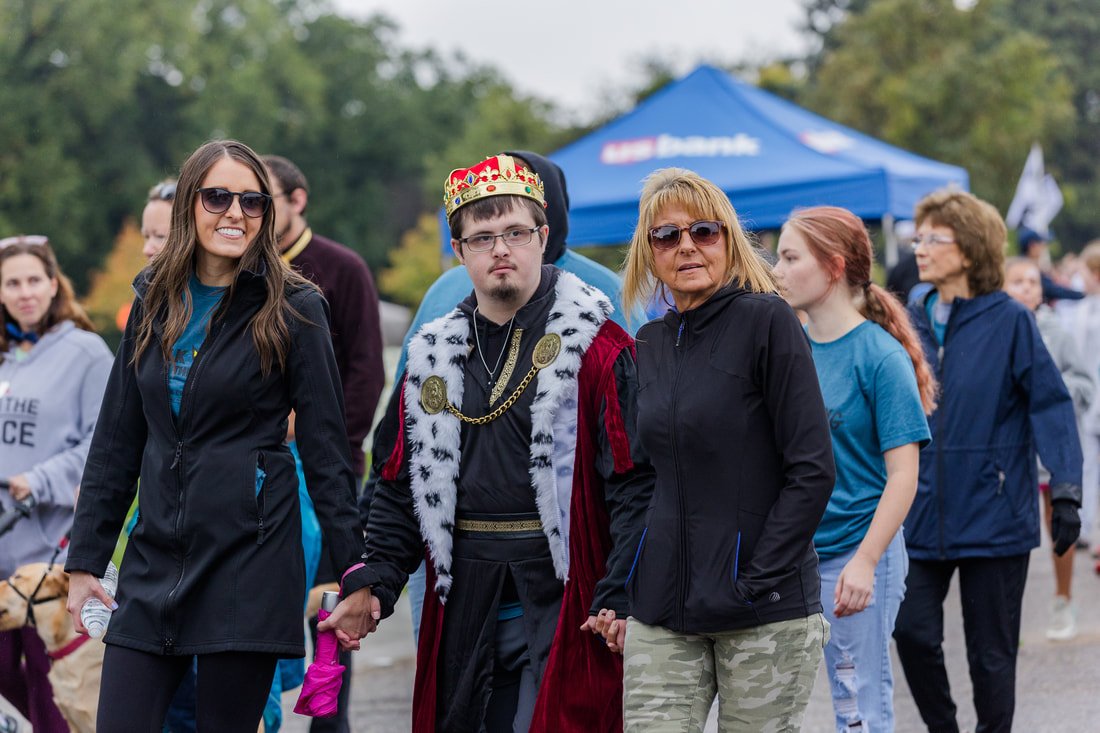 Group of people walking outdoors during daytime event, some wearing casual clothes, and a man dressed as a king or monarch with a crown, robes, and medals.