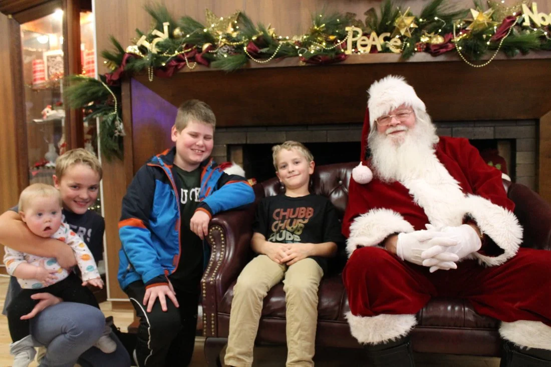 A group of five children and Santa Claus sitting in front of a decorated fireplace with festive greenery and ornaments, celebrating Christmas.
