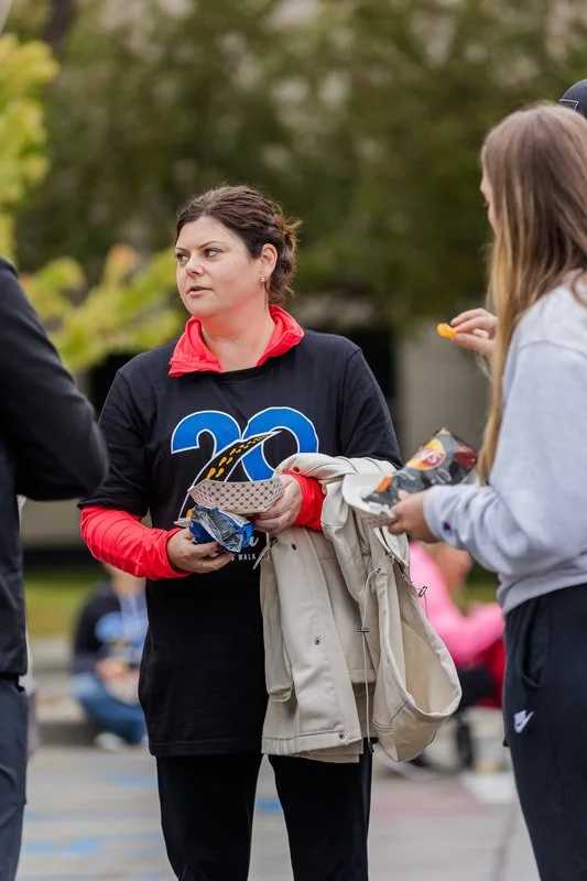 A woman in a black T-shirt with blue and white graphics, a red hoodie underneath, and black pants, holding a beige coat and food, talking to two other women outdoors.