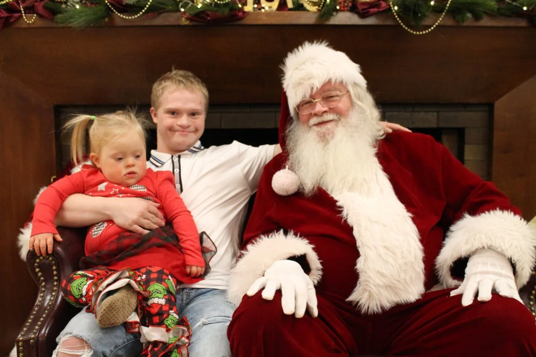 A young girl sitting on Santa Claus's lap, with an older boy standing beside them, all smiling indoors decorated with Christmas garlands.