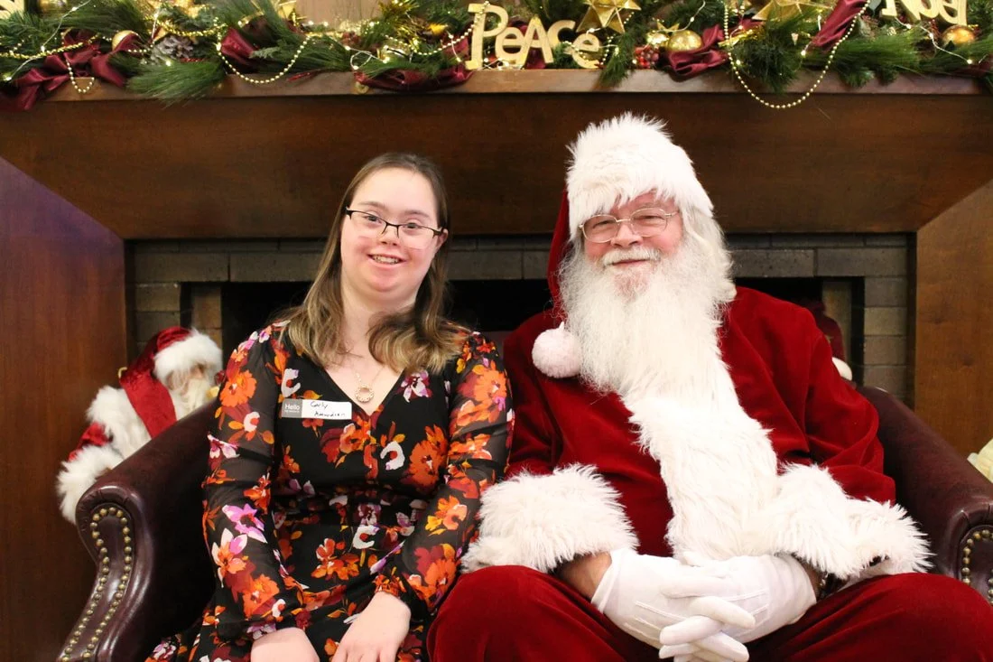 A young woman in a floral dress sitting next to Santa Claus in a red suit, in front of a fireplace decorated for Christmas with garlands, ornaments, and a 'Peace' sign.
