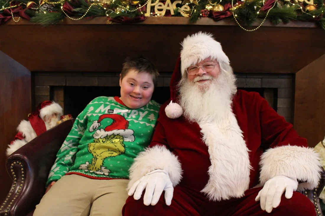 A boy smiling and sitting next to a man dressed as Santa Claus, both sitting in front of a decorated fireplace with Christmas garland and ornaments.