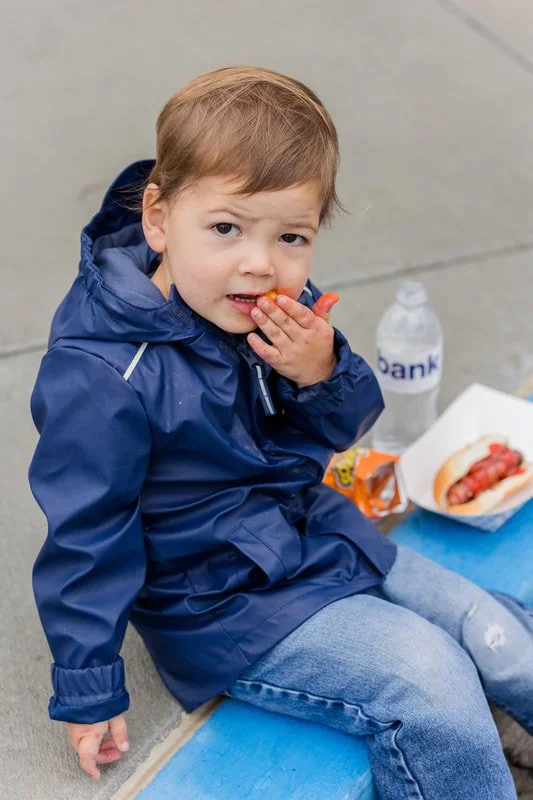 A young boy with light brown hair and wearing a blue rain jacket and jeans sits on a concrete surface, eating hot dog and chips with a bottle of water nearby.