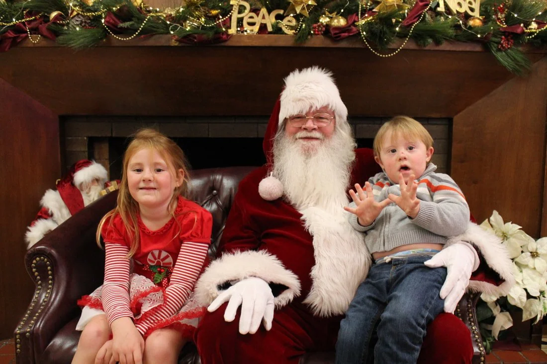 A man dressed as Santa Claus sitting on a brown leather couch with two children, in front of a decorated fireplace mantel with Christmas ornaments and greenery.