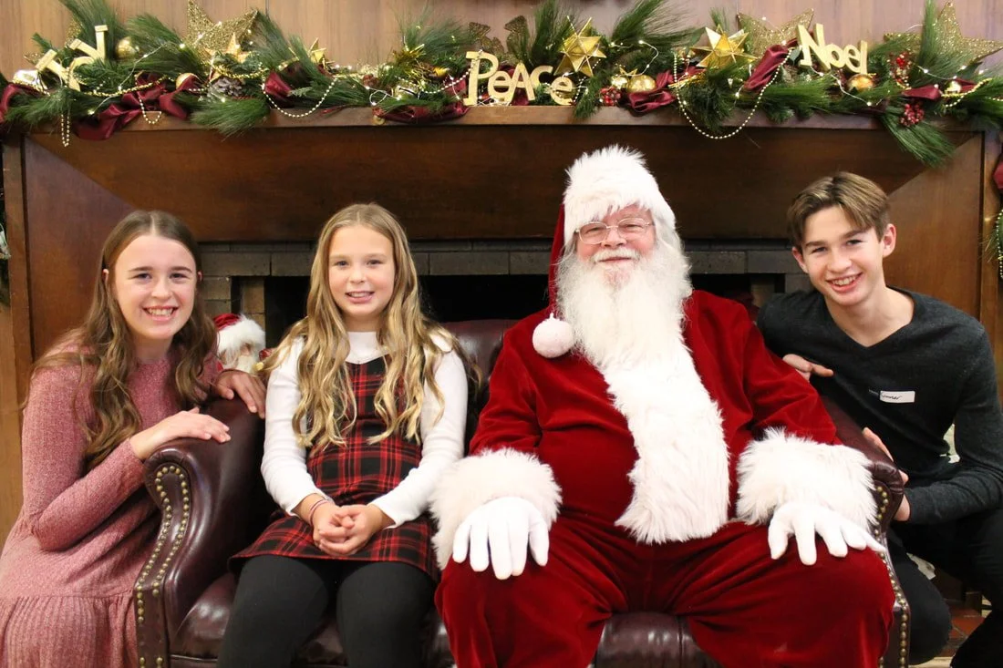 Three children with Santa Claus sitting in front of a decorated fireplace mantle with Christmas garland and signs reading 'Peace', 'Noel' and 'Joy'.
