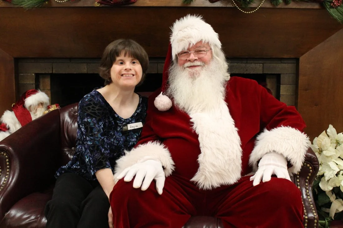 A young girl sitting next to Santa Claus, both smiling, inside a decorated room with a fireplace and Christmas decorations.