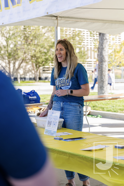 A smiling woman standing at an outdoor booth under a white canopy, engaging with visitors at a daytime event, with trees and people in the background.