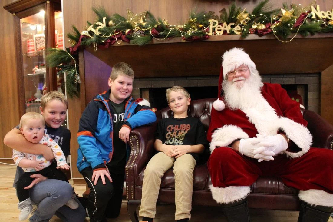 Four children and Santa Claus sitting on a leather couch in a room decorated for Christmas with a fireplace, holiday garland, and ornaments.