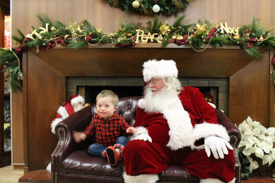 A young boy sitting next to Santa Claus on a leather couch in a decorated room.