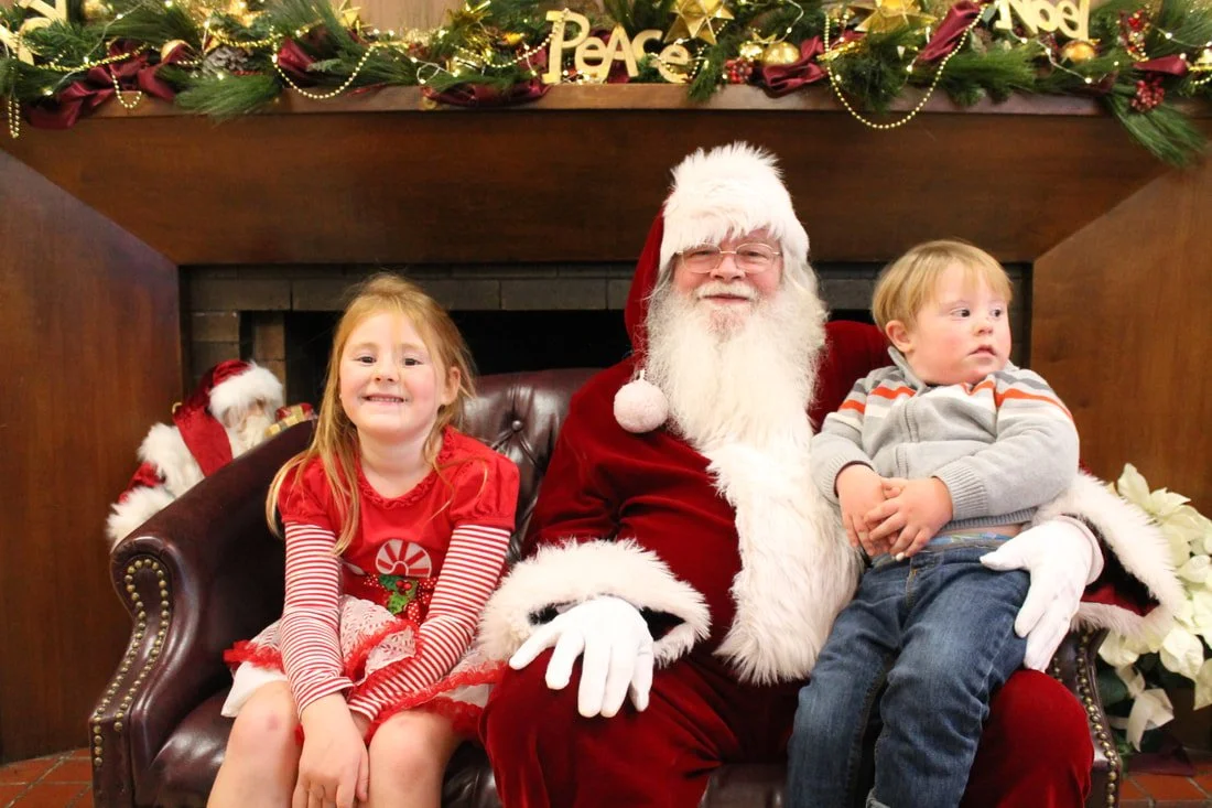 Smiling young girl in a red festive dress sitting on Santa Claus's lap, with a young boy sitting next to Santa, in a Christmas decorated setting with garland and 