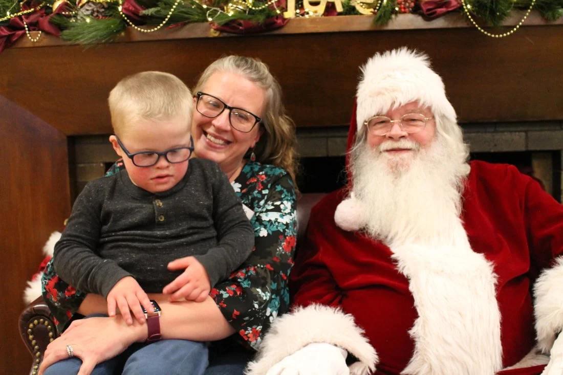 A woman with glasses holding a young boy with glasses, sitting next to a man dressed as Santa Claus. The scene is festive with Christmas decorations and lights in the background.