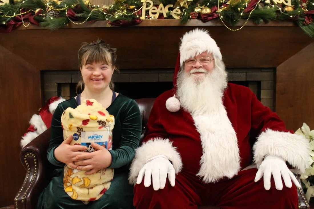 A girl smiling and holding a blanket with Mickey Mouse design, sitting next to a man dressed as Santa Claus, in front of a decorated fireplace mantle with Christmas decorations.