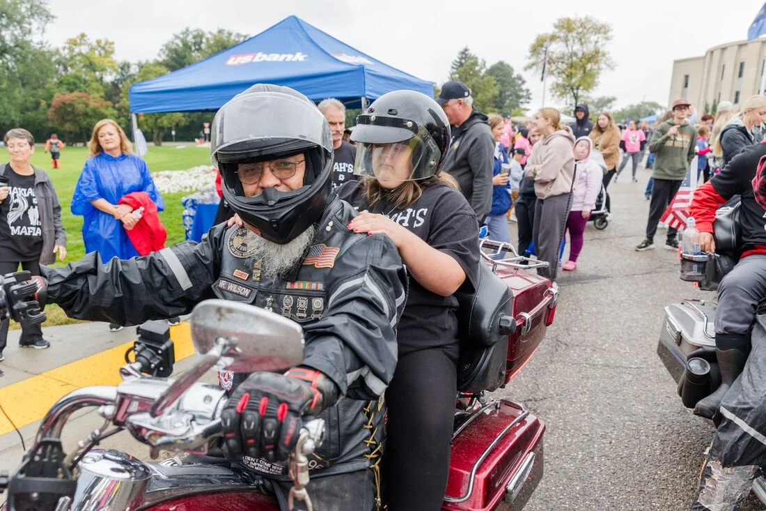 A man and woman riding a motorcycle at an outdoor event with a crowd of people in the background, some holding American flags and wearing casual clothing.