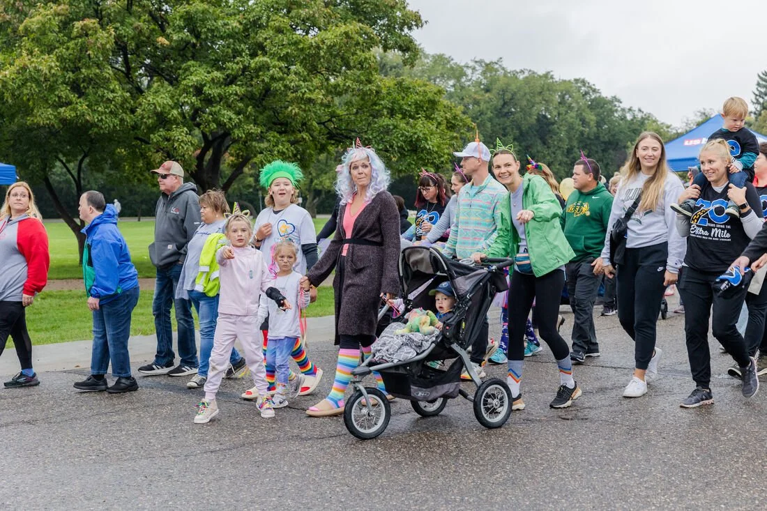People participating in a fun walk or parade, some dressed in colorful costumes and accessories, in an outdoor park setting with trees and cloudy sky.