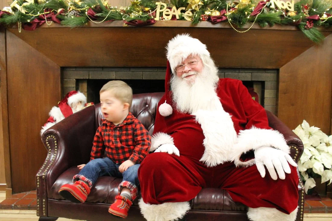 A young boy sitting next to Santa Claus on a leather couch in front of a fireplace decorated with holiday garland and ornaments, some figurines of Santa Claus in the background, and poinsettia flowers nearby.