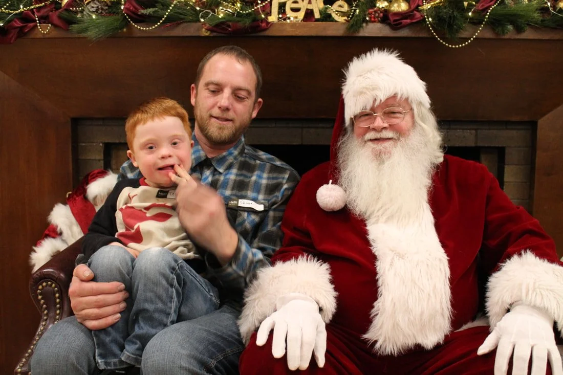 A man, a young boy, and Santa Claus sitting together in front of a fireplace decorated with holiday garland and ornaments.