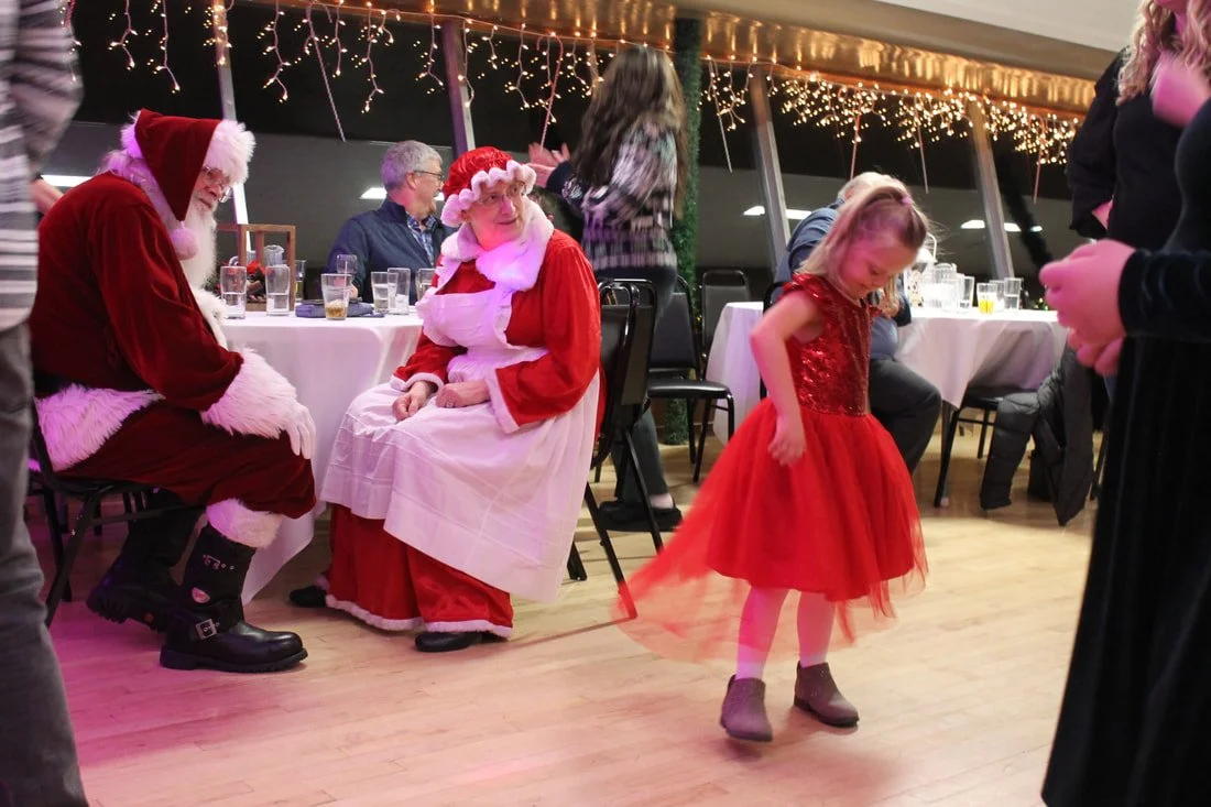 A Christmas celebration with Santa and Mrs. Claus sitting at a table, while a young girl in a red dress dances in front of them. Festive string lights decorate the background.