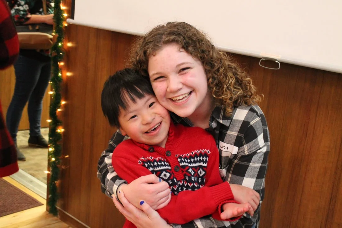 A young woman with curly hair hugging a young boy with straight black hair, both smiling. The woman has a name tag that says 'Lucy'.