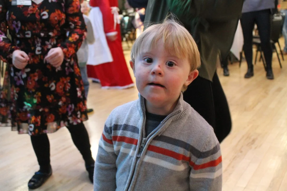 Young boy with blond hair and freckles looking at the camera during a festive indoor event with others in the background.