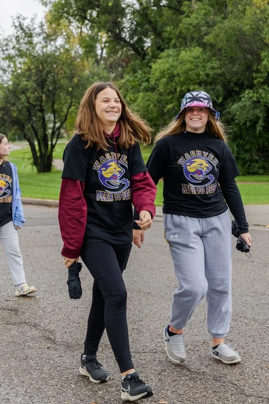 Two teenage girls walking outdoors, smiling, wearing matching black t-shirts with a dragon logo and text, with one girl wearing a bucket hat and grey sweatpants.