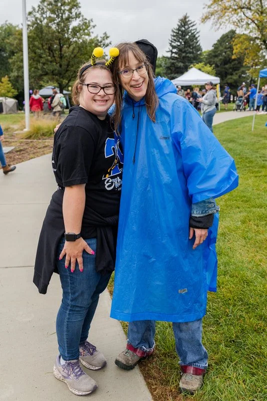 Two smiling women standing outdoors on a cloudy day, one wearing a black shirt with a blue logo, bee antenna headband, and jeans, the other wearing a blue raincoat, glasses, and jeans, with a crowd of people and tents in the background.