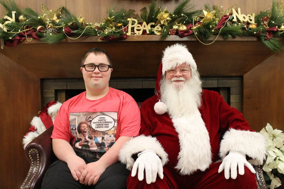 A boy with glasses sitting next to Santa Claus, both smiling, in front of a fireplace decorated with Christmas garland and ornaments. The garland has gold stars, red ribbons, and the words "Peace" and "Noel".