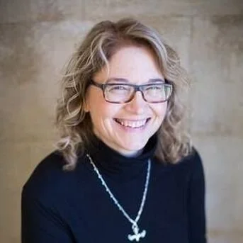 A smiling woman with glasses, wavy blonde hair, wearing a black turtleneck and a shell necklace.