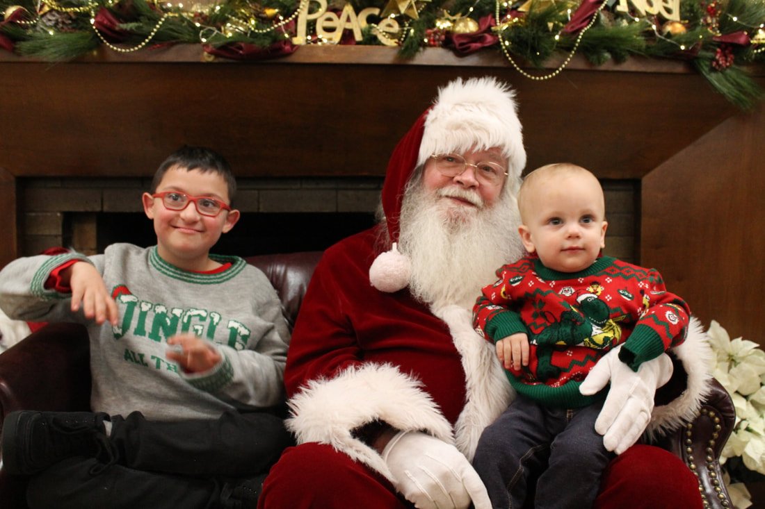 Two young children sitting with Santa Claus, all dressed in Christmas sweaters, in front of a fireplace decorated with garland and holiday ornaments.